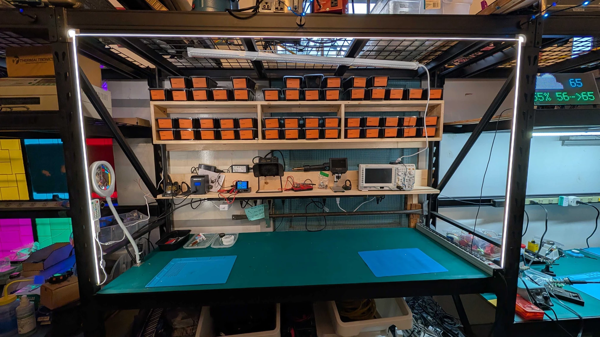 An electronics workbench with new strip lighting, orange-labeled storage bins, test instruments, and two soldering mats